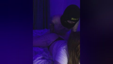 Video of a man in a black cap with \"Boon\" text, lying on a bed with white sheets, under purple lighting.