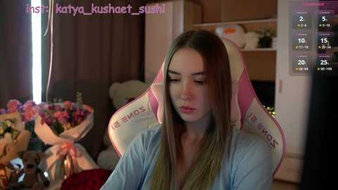 Media: A video of a young woman with long brown hair, wearing a blue hoodie, sitting in a pink gaming chair in a cozy, dimly lit room with a TV and floral arrangement.