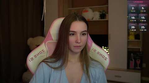 Media: Video of a young woman with long brown hair, wearing a pink gaming chair and a blue shirt, in a dimly lit room with a white shelf in the background.