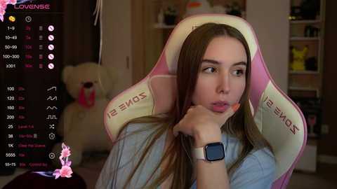 Media: A video of a young woman with long brown hair in a pink gaming chair, wearing a light blue shirt and a smartwatch, looking thoughtful.