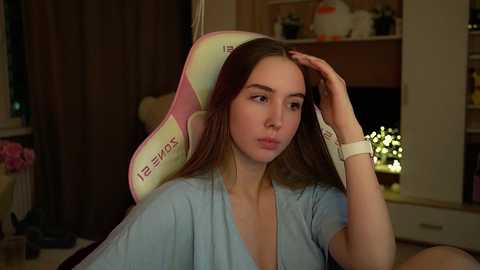 Media: Video of a young Caucasian woman with light skin and straight brown hair, sitting in a blue hospital gown, looking tired with hand on head, in a dimly lit room with shelves and a window.