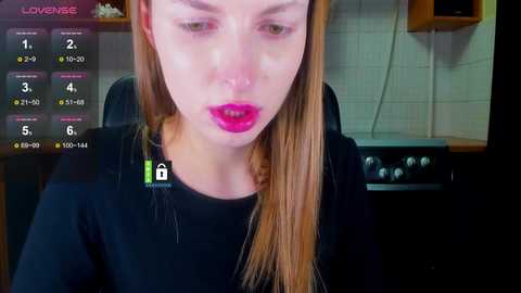 Video of a young woman with long, straight blonde hair, wearing bright pink lipstick, seated in a kitchen with tiled walls and a stove.