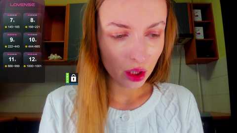 Video of a woman with long, straight, light brown hair and fair skin, wearing a white blouse. She has a sad expression. Background shows a kitchen with wooden cabinets and a TV screen displaying a game.