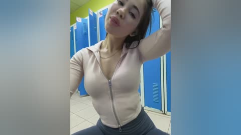 Video of an Asian woman with long black hair, wearing a light pink zip-up hoodie and high-waisted dark leggings, standing in a brightly lit, blue-locked restroom with tiled floors.