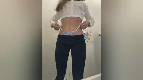 Media: Video of a slender woman in a white shirt, exposing blue floral-patterned panties, standing in a narrow, beige-walled bathroom with a colorful sticker on the wall.