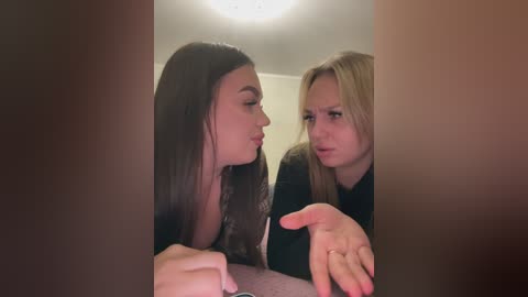 Video of two young women with long hair, one brunette and one blonde, discussing something intently. They are indoors with a ceiling light overhead.