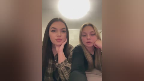 Video of two young women with fair skin, long dark hair, and plaid shirts, sitting close together indoors. They have their heads propped up on their hands, looking relaxed and smiling.