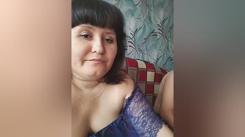 Video of a middle-aged woman with short black hair and a blue lace off-shoulder top, smiling subtly. Background features a teal and brown patterned wall and a red and beige checkered cushion.