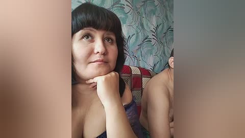 Video of a middle-aged woman with short dark hair and light skin, wearing a black strapless top, resting her chin on her hand, against a background of blue and green floral wallpaper.