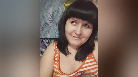 A video of a smiling, middle-aged woman with shoulder-length black hair, wearing an orange and white striped tank top, seated indoors with floral wallpaper and a metal chair visible in the background.