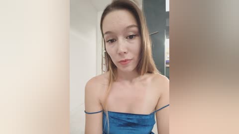Media: A video of a young, fair-skinned woman with long brown hair, wearing a blue velvet off-shoulder dress, standing indoors with white and gray walls in the background.