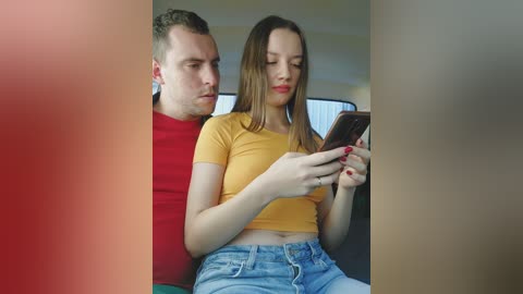 Video of a young couple in a car: a blonde woman in a mustard crop top, denim shorts, and red nails, holding a phone, next to a man in a red shirt.