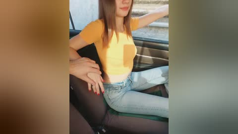 Video of a young woman with straight brown hair, wearing a tight orange crop top and light blue jeans, seated in a car with a man's hand on her thigh.