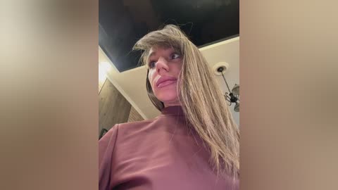 A video of a blonde woman in a pink turtleneck sweater, with a dark room and ceiling light visible in the background.