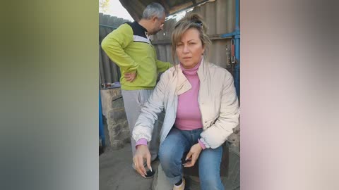 Media: Video of a middle-aged blonde woman in a beige jacket and pink turtleneck, sitting on a wooden bench beside an older man in a green jacket, in a rustic, outdoor setting with metal fencing and a corrugated roof.