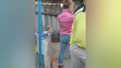 Media: Video of two women standing at a stone counter in a rustic outdoor kitchen. One wears a pink jacket and blue jeans, the other a green jacket and gray pants. Blue and yellow poles frame the scene.