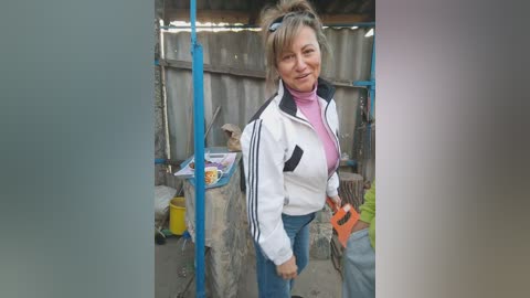 Media: Video of a smiling middle-aged woman with light brown hair in a ponytail, wearing a white jacket, pink turtleneck, and blue jeans, standing in a cluttered, makeshift outdoor workshop.