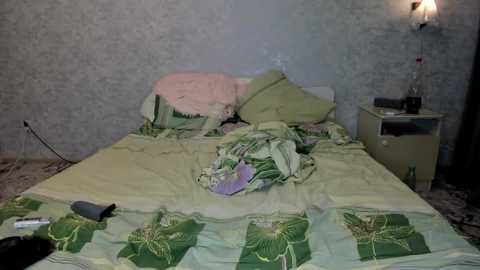 A video of a messy bedroom with a bed covered in disheveled green and yellow bedding, a pink pillow, and scattered clothes. A beige nightstand holds a water bottle and a black object.