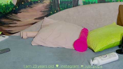 Media: A video shows a cozy, cluttered bed with a pink pillow, a green blanket, a bottle, and a wooden nightstand against a forest mural.