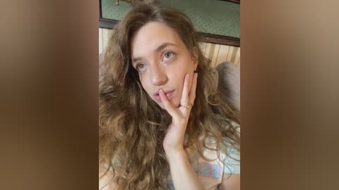 A video of a fair-skinned woman with long, curly brown hair, wearing a white t-shirt, sitting with her hand on her cheek, set in a cozy, wooden-paneled room.