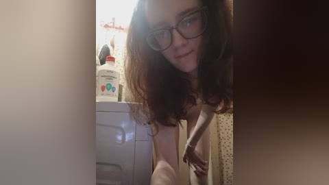 A video of a fair-skinned woman with long brown hair, wearing glasses and a white shirt, leaning over a washing machine in a dimly lit bathroom.