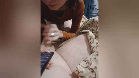 A video showing a woman with light skin, brown hair, and a brown sweater, leaning over a man's chest with a remote in hand, while he lies on a bed with patterned sheets.