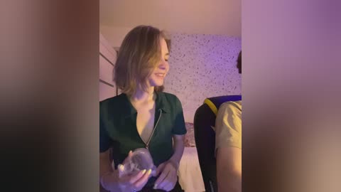 A video of a smiling woman with light brown hair, wearing a dark green, unzipped top, holding a baby, in a dimly lit room with a speckled wallpaper and a bed.
