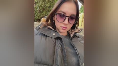 Media: Video of a young woman with light skin, straight brown hair, and large round sunglasses, wearing a dark puffy jacket, standing outdoors in front of green foliage.