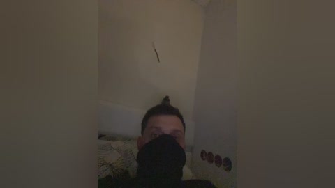 A video captures a person with dark hair, partially obscured by a black mask, standing in a dimly lit, narrow hallway with white walls and a patterned rug.