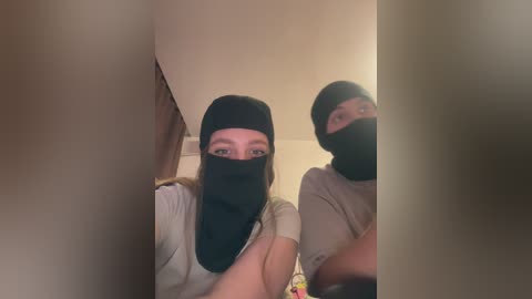 Video of two women with fair skin, wearing black masks and beige t-shirts, standing in a dimly lit room with beige walls and a mirror reflecting their faces.