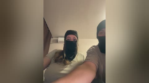 A video of two masked individuals in a dimly lit room with beige walls and a ceiling fan. Both wear black masks and light-colored shirts, suggesting a covert or clandestine activity.