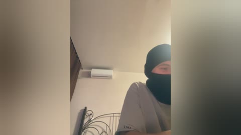 A video of a person wearing a black balaclava, gray T-shirt, and shorts standing in a small, dimly lit room with beige walls, an air conditioner, and a decorative metal frame.