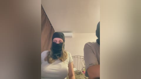 Video of a woman with long blonde hair and a black face mask, standing in a dimly lit bedroom with beige walls, a metal bed frame, and a white AC unit.