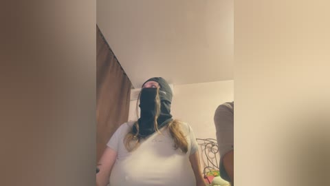 Video of a woman with fair skin, wearing a black ski mask, white t-shirt, and braided hair, standing in a dimly lit room with beige walls and a wooden door.
