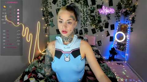 Video of a tattooed, light-skinned woman in a blue bunny costume, with a gray background, fairy lights, and a neon sign reading \"vibes.\