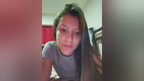 Video of an Asian woman with long, straight, dark hair, wearing a gray T-shirt, sitting on a bed in a dimly lit room with red curtains and a wooden headboard.