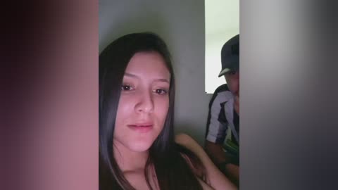 Video of an Asian woman with straight black hair, wearing a black top, smiling slightly. She is partially obscured by a vertical light-colored panel on the left and a blurred reflection of a boy in a baseball cap on the right.