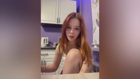 Video of a young Caucasian woman with long, reddish-brown hair, sitting on a kitchen counter, wearing a strapless top, in a modern kitchen with white cabinets and purple walls.