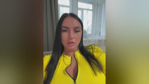 Video of a middle-aged woman with long black hair, wearing a bright yellow jacket, seated indoors near a window with white curtains.