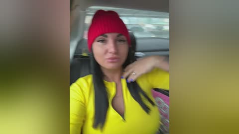 A video of a woman with long black hair and a red beanie, wearing a bright yellow long-sleeve shirt, seated in a car with blurred surroundings.