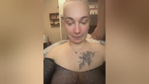 Video of a fair-skinned woman with short blonde hair, wearing a sequined black dress, showcasing a large floral tattoo on her chest. She is in a dimly lit room with beige walls, shelves, and a door.