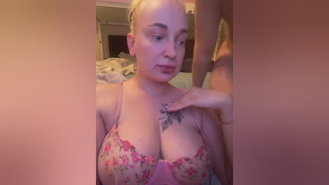 Video of a light-skinned, busty woman with short blonde hair, wearing a pink floral bra, in a bedroom. A man's hand is placed on her chest, revealing a tattoo.