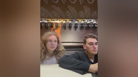Media: Video of two young people, a girl with curly blonde hair and a boy with short brown hair, sitting in a modern, dimly lit restaurant with brown walls and white, repetitive patterns. They appear engrossed in conversation.