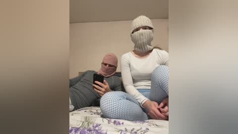 Video of a woman in a white long-sleeve shirt and blue leggings, wearing a gray knitted mask, sitting on a bed with a man in a gray hoodie and pink mask. Both are focused on a smartphone.