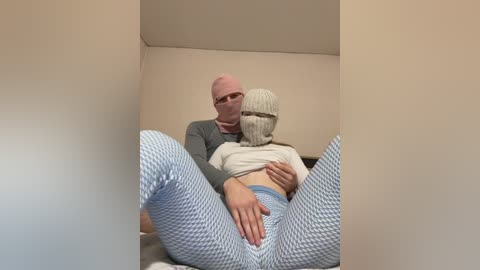 Video of a man in a grey sweater and woman in a white top and blue patterned leggings, seated in a corner of a beige room, with a white mask covering the woman's face.
