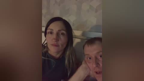 Video of a woman with long, straight hair and a man with short hair, both looking into the camera, in a dimly lit room with a wallpaper pattern in the background.
