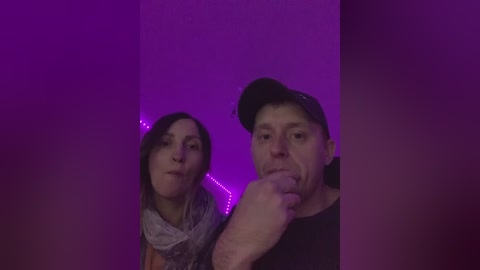 Video of a woman with light skin and medium-length brown hair, wearing a gray scarf, and a man with light skin, wearing a black cap and black shirt, against a purple-lit background.