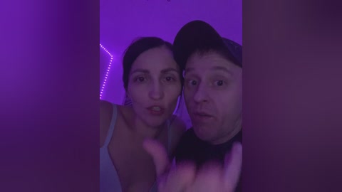 Video of two people, a woman and a man, standing close together, illuminated by purple lighting. The woman has dark hair, wears a light-colored top, and looks slightly to the left. The man has a light complexion, wears a baseball cap, and looks directly at the camera.