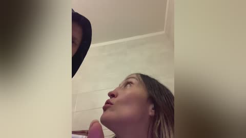 A video shows a young woman with long, wet hair, looking upward in a bathroom with beige tiles, a mirror, and a pink object in the foreground.