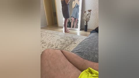 Media: Video of a man in a gray shirt and yellow shorts, standing in a room with beige walls and a wooden floor, holding a mirror to see his reflection.